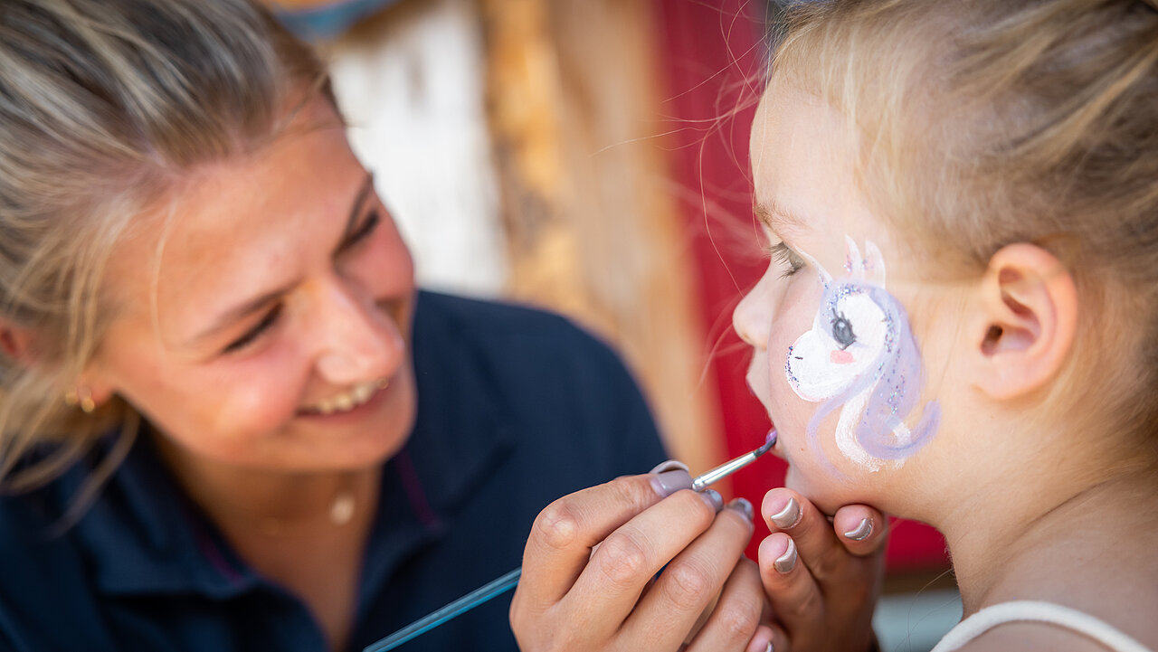 Animationsmitarbeiterin malt mit Kinderschminke auf Wange eines Mädchens Animationsmitarbeiterin malt mit Kinderschminke auf Wange eines Mädchens